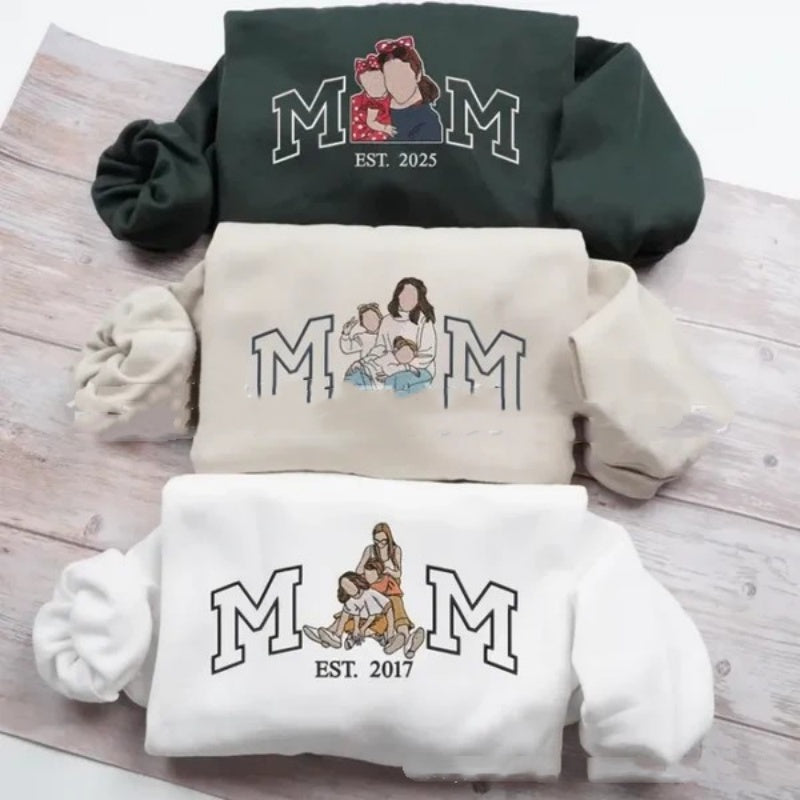 Three sweatshirts with 'MOM' text and illustrations on a wooden surface